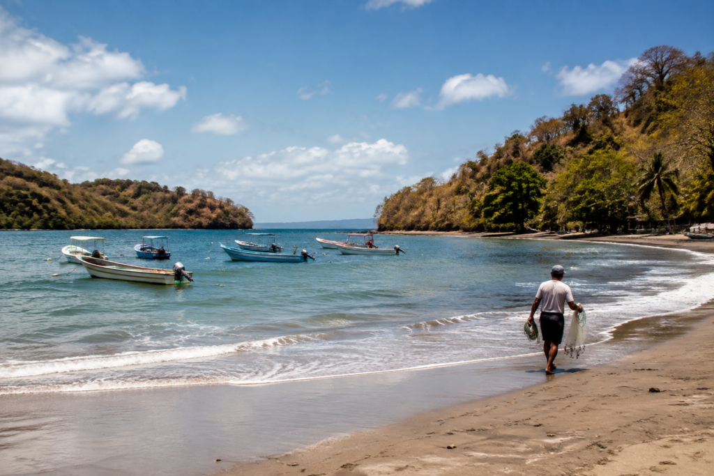 Small boats float in a tropical bay bordered by a sandy beach and wooded hills.