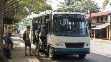 Passengers board a bus beside a shaded street with low-rise buildings.